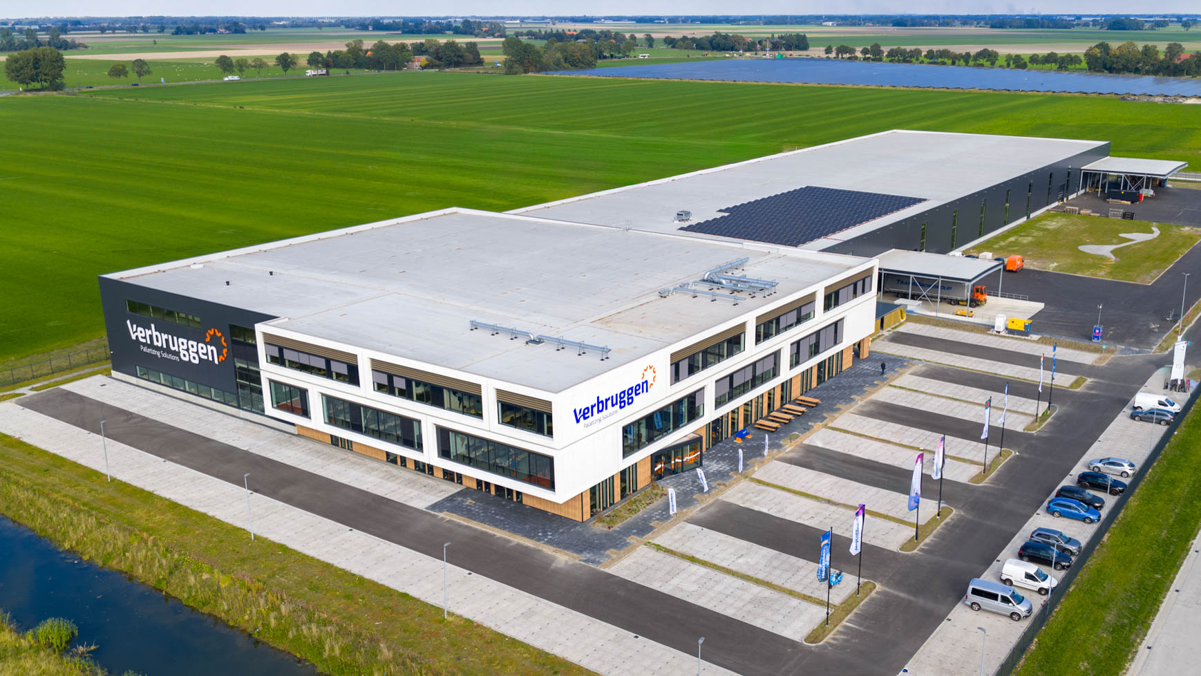 Aerial view of the Verbruggen Palletizing Solutions headquarters — a modern two-story office and warehouse building with a dark and white facade, rooftop solar panels, a parking lot with cars and event flags, set among flat green fields and canals under an overcast sky.